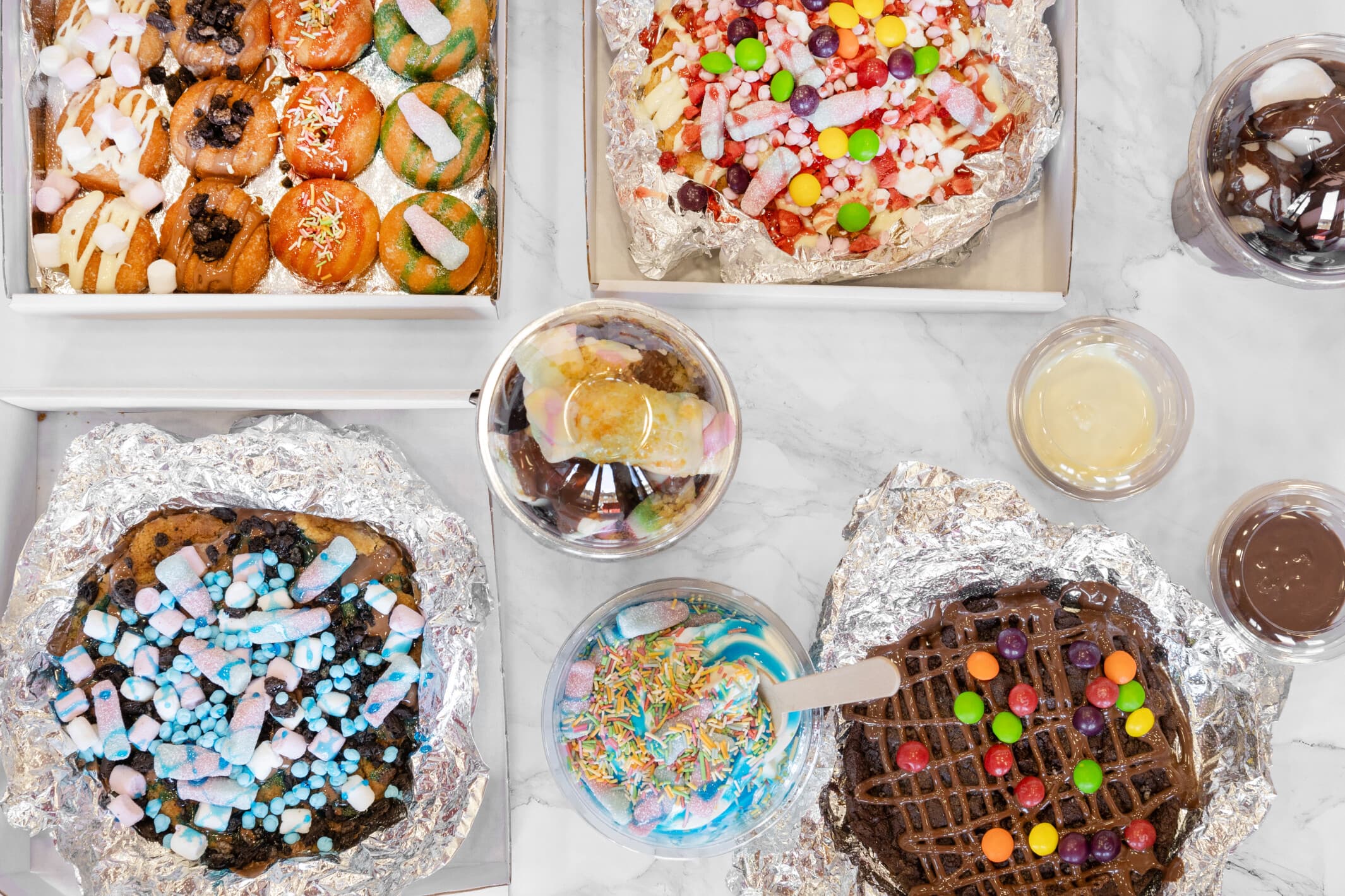 An overhead spread of ShakeTastic loaded cookie dough, doughnuts and dessert cups covered in sweets and sauces
